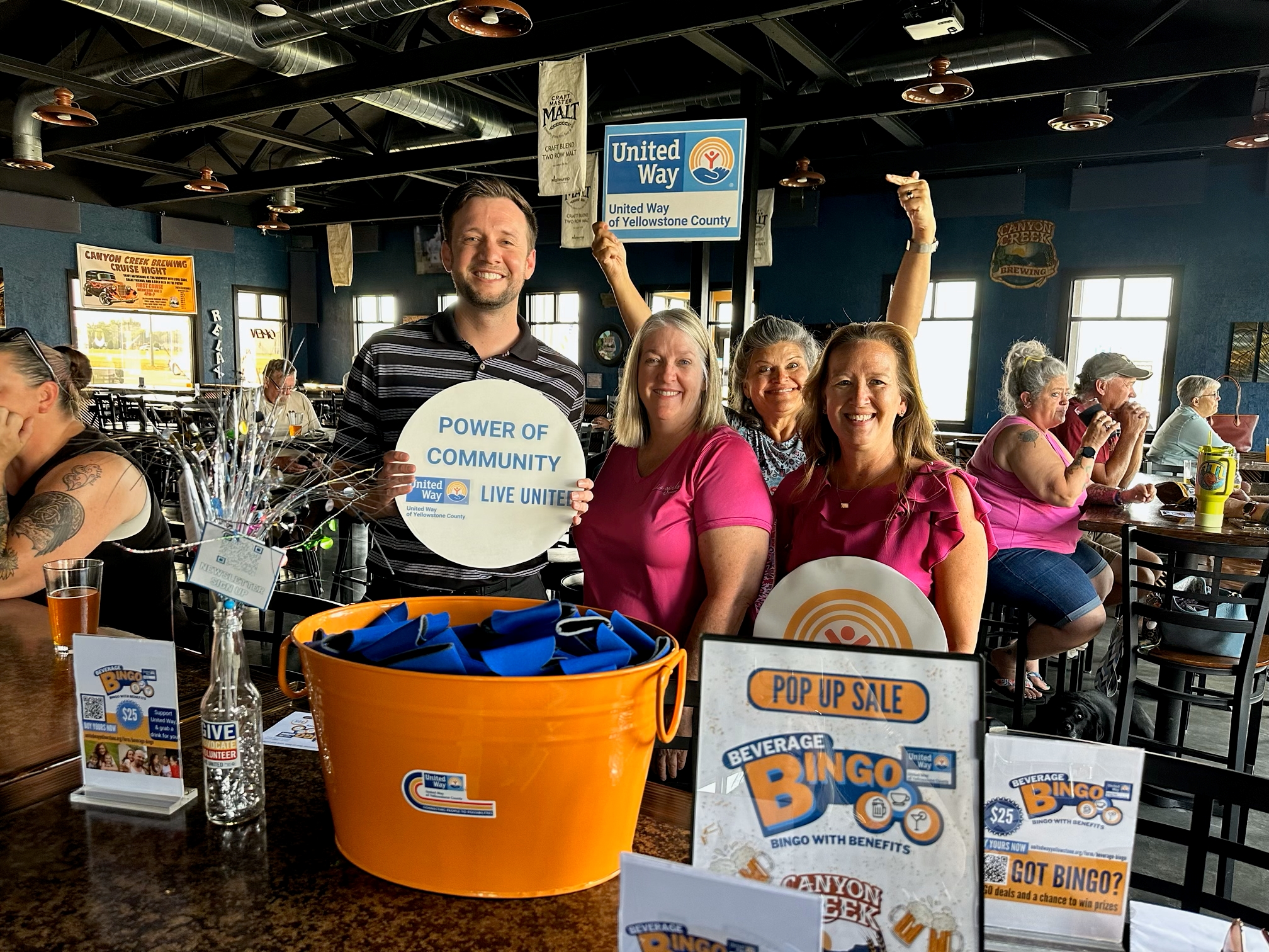 Board member Nathan Hirsch holds a United way sign at Canyon Creek Brewing in a group of three other people at a Beverage Bingo event. 